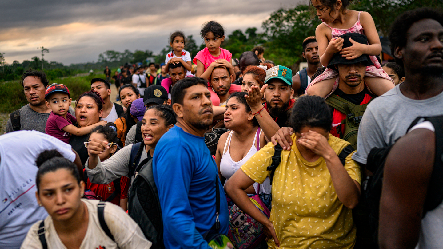 photo of crowded line of people wearing backpacks, several carrying a child on their shoulders, one woman with hand to forehead