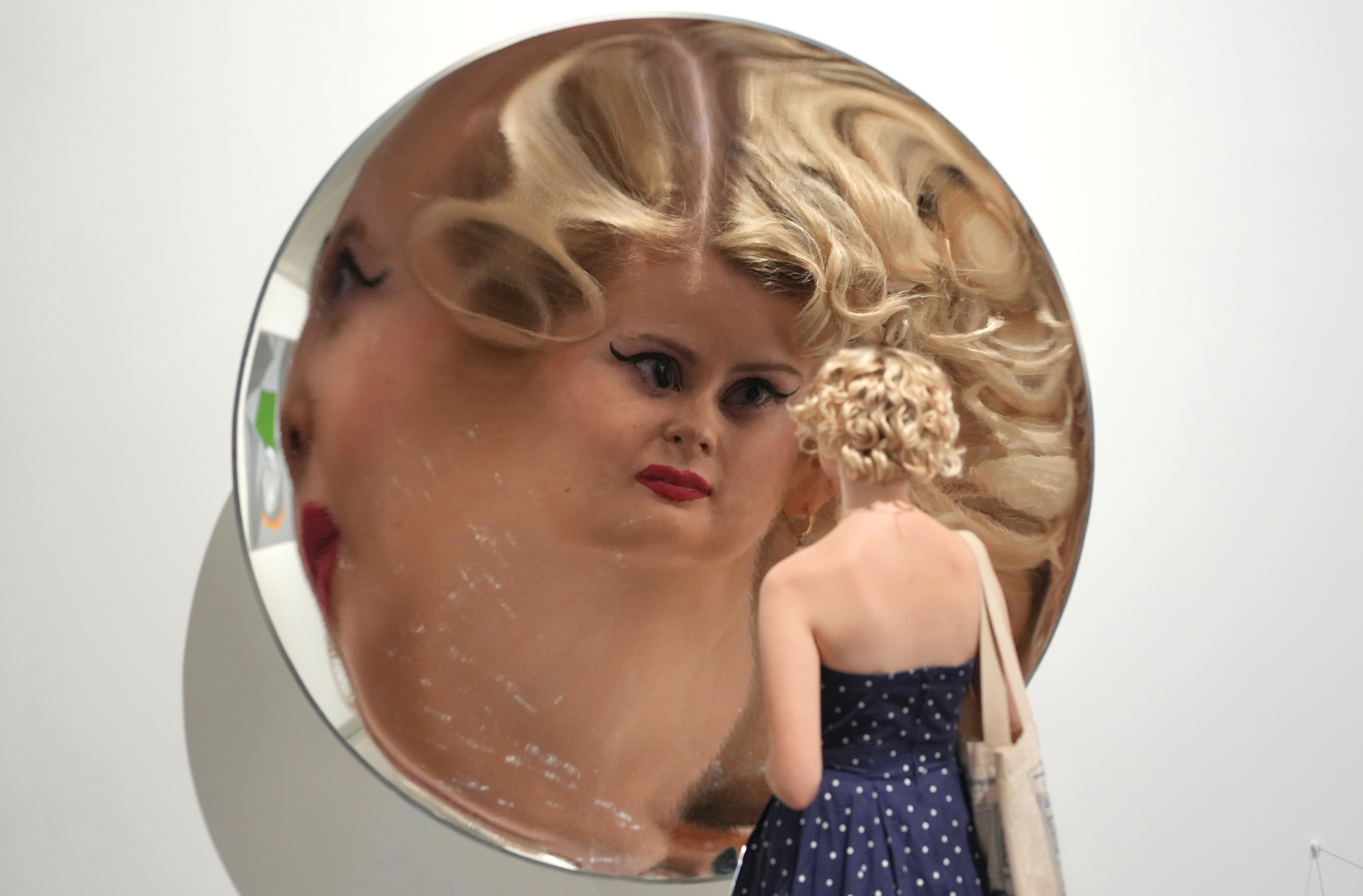 A person stands close to a huge circular curved mirror, their face magnified and distorted in the reflection.