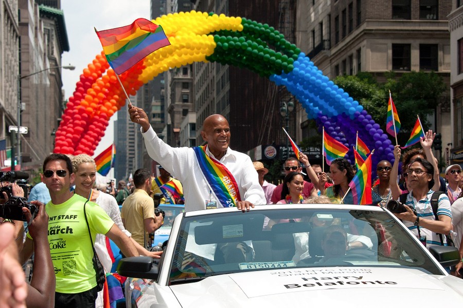 Harry Belafonte waves a rainbow flag while riding in an open convertible in a parade.